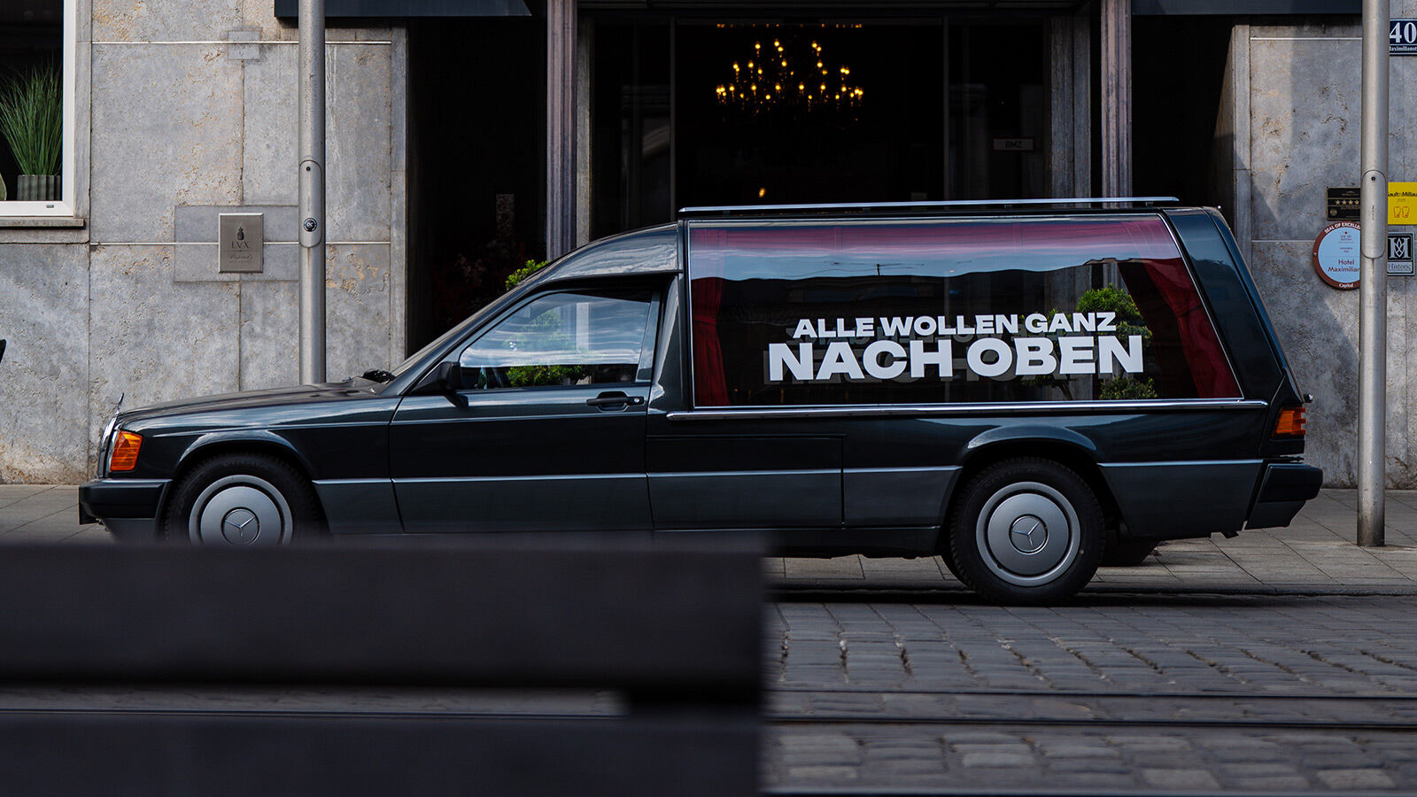 Der beklebte Leichenwagen in ganzer Länge, geparkt vor einer Häuserfront in der Innenstadt.