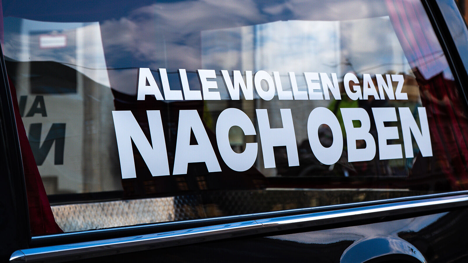 Heckscheibe eines Leichenwagens mit dem weißen Schriftzug „Alle wollen ganz nach oben“.