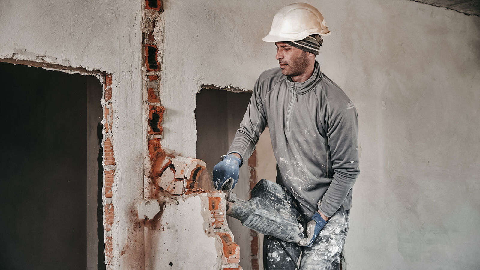 Ein Bauarbeiter mit Helm öffnet mit dem Abbruchhammer eine gemauerte Wand.