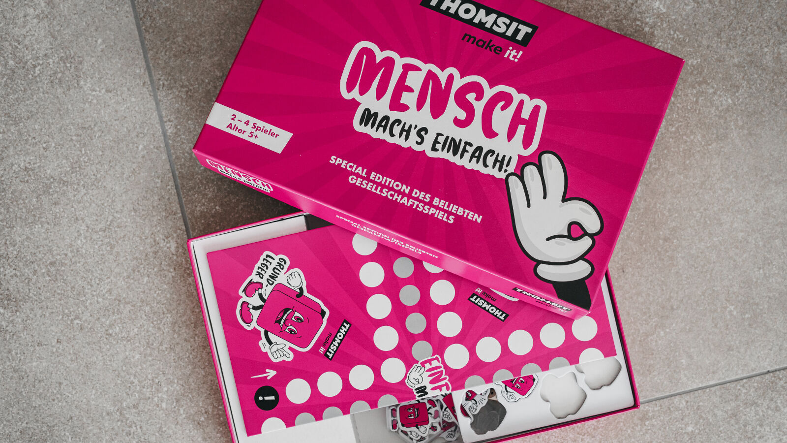 THOMSIT-Sonderedition des Spiels Mensch ärgere dich nicht: pinke Schachtel „Mensch mach’s einfach!“ neben dem Spielfeld.