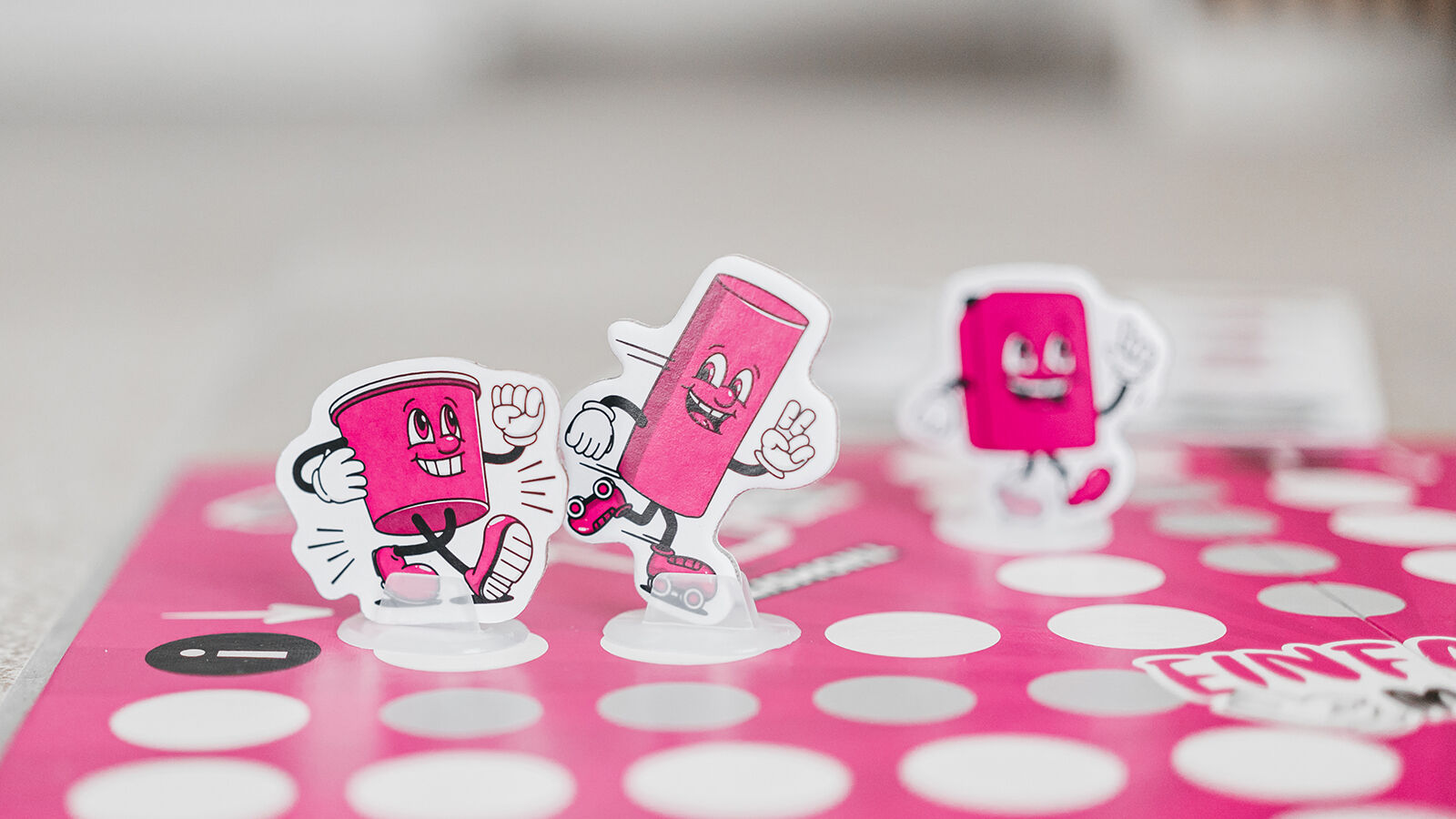 Spielfiguren der THOMSIT-Sonderedition: Eimer und Kartusche als Comicfiguren auf dem pinken Spielfeld.