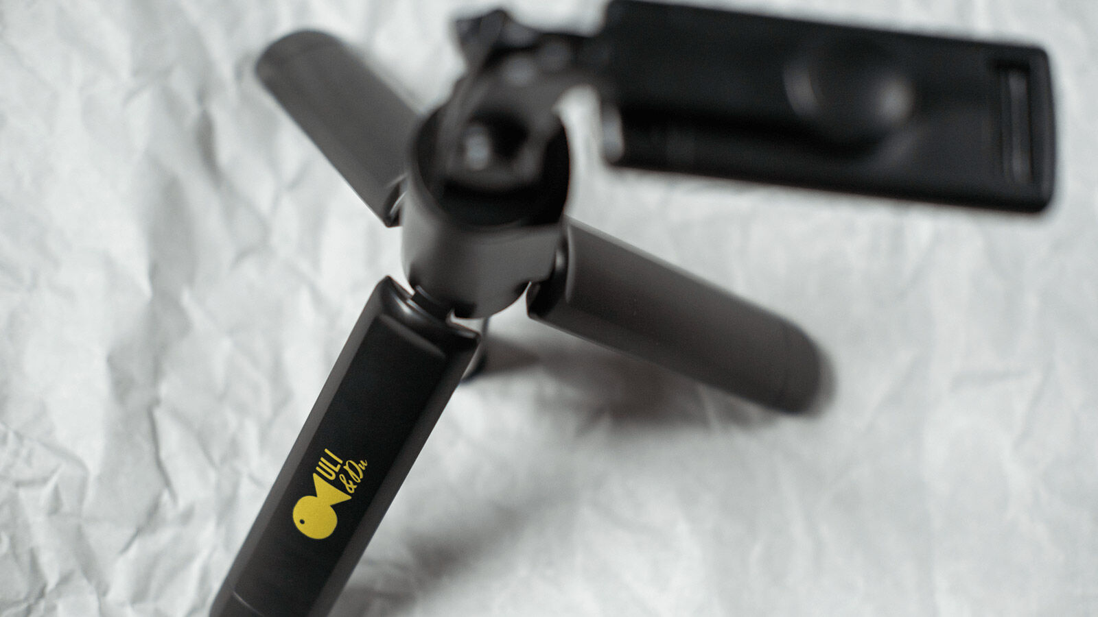 Schwarzer Selfiestick mit dem gelben Uli-&-Du-Logo, daneben die Handyhalterung.